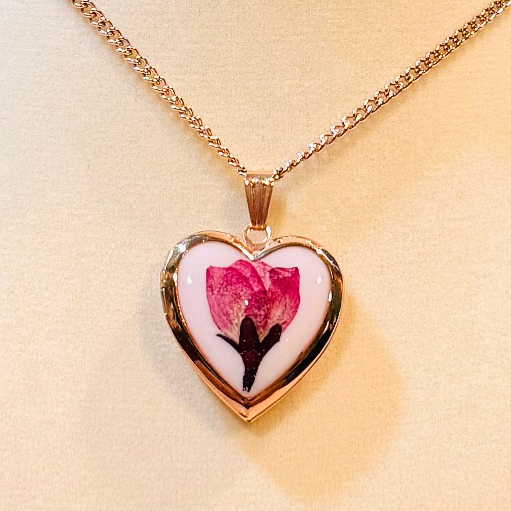 Heart-shaped pendant with a pink flower design on a gold chain against a beige background